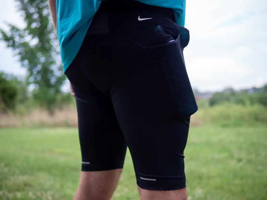 A runner wearing the half tights, with a 500ml soft flask in the side pocket and a 150ml nutrition flask in the front right waist pocket, shown from a rear side angle