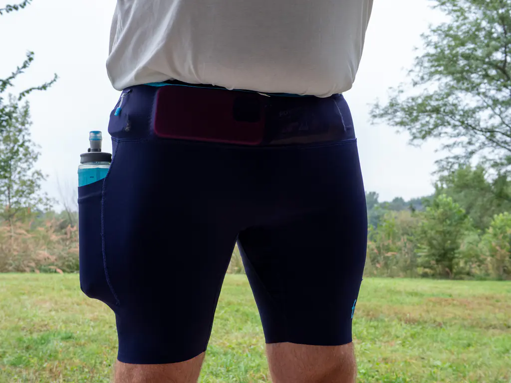 A front view of a runner wearing the half tights, with an iPhone 16 Pro stowed in the front waistband storage and a 500ml soft flask in the thigh pocket